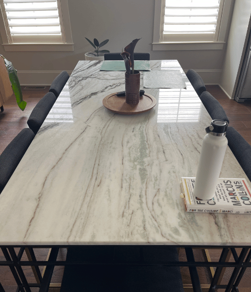 Marble dining table with black metal legs and gray upholstered chairs; a potted plant on a wooden tray at the center, with a water bottle and books on the right side.